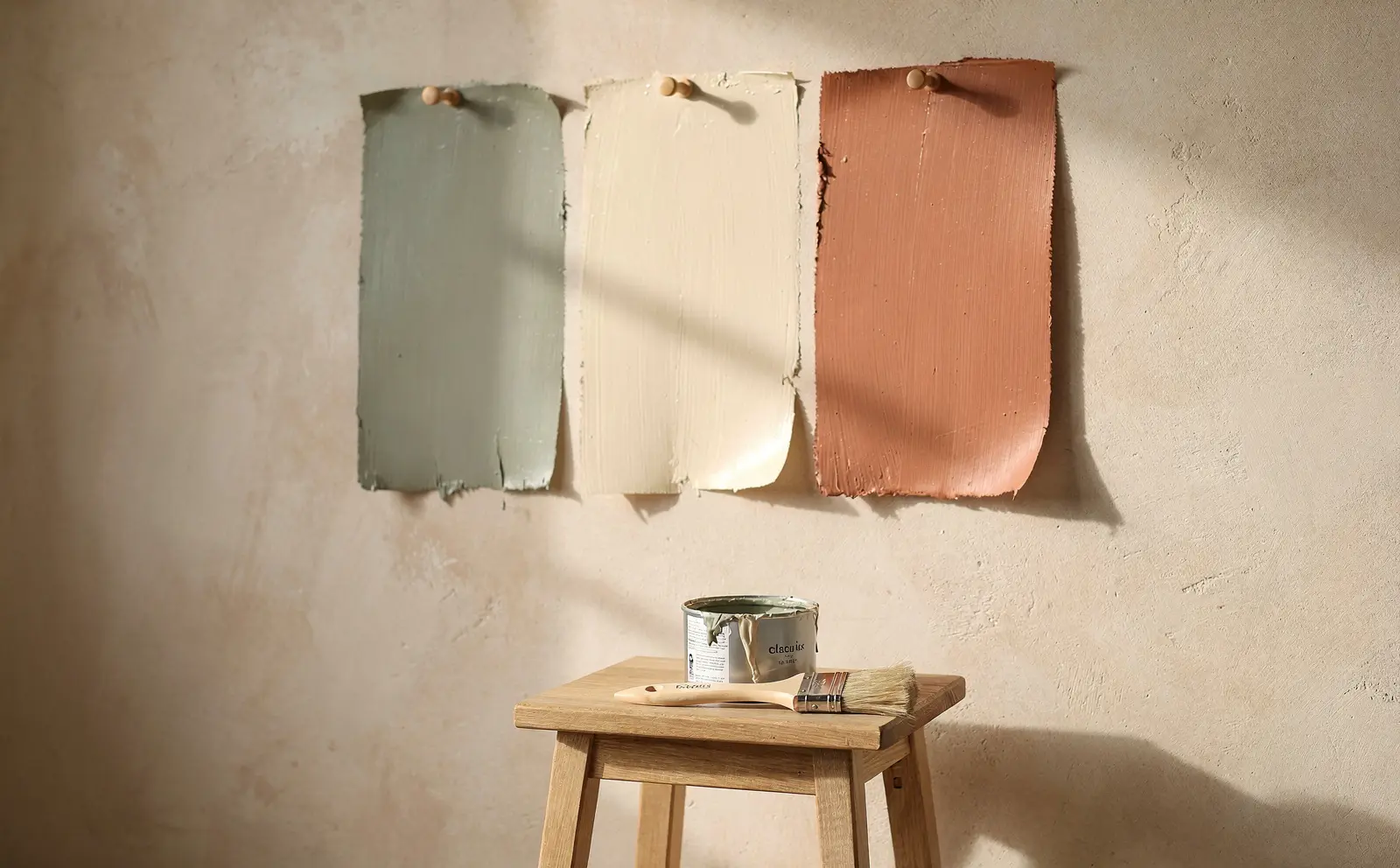 Sage, cream and clay paint swatches pinned on a plaster wall above a paint tin on an oak stool